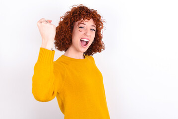 Overjoyed young redhead girl wearing yellow sweater over white background glad to receive good news, clenching fist and making winning gesture.