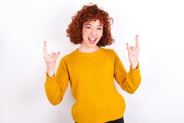 Born to rock this world. Joyful young redhead girl wearing yellow sweater over white background screaming out loud and showing with raised arms horns or rock gesture.