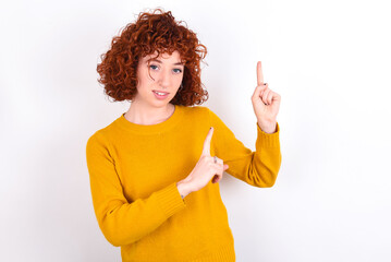 Positive young redhead girl wearing yellow sweater over white background with beaming smile pointing with two fingers and looking on empty copy space. Advertisement concept.