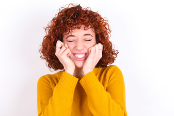 Fototapeta na wymiar Portrait of young redhead girl wearing yellow sweater over white background being overwhelmed, expressing excitement and happiness with closed eyes and hands near face.