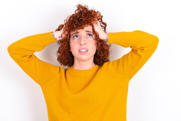 Horrible, stress, shock. Portrait emotional crazy young redhead girl wearing yellow sweater over white background clasping head in hands. Emotions, facial expression concept.