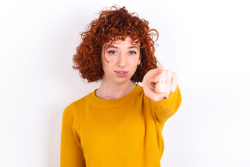 Cheerful young redhead girl wearing yellow sweater over white background indicates happily at you, chooses to compete, has positive expression, makes choice.