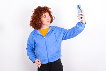 Portrait of a young redhead girl wearing blue jacket over white background  taking a selfie to send it to friends and followers or post it on his social media.