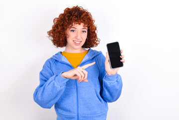 Smiling young redhead girl wearing blue jacket over white background showing and pointing at empty phone screen. Advertisement and communication concept.