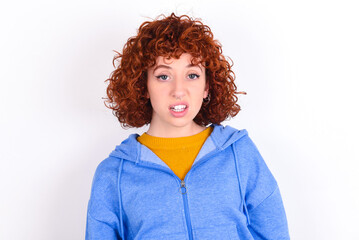 Portrait of dissatisfied young redhead girl wearing blue jacket over white background smirks face, purses lips and looks with annoyance at camera, discontent hearing something unpleasant