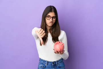 Young caucasian woman holding a piggybank isolated on purple background showing thumb down with...