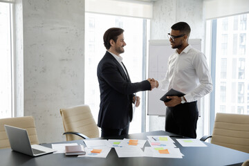 Smiling young male team leader shaking hands with African American male employee, praising for good job. Happy two diverse businessmen making agreement or establishing partnership, cooperation concept