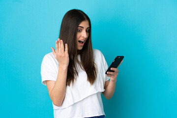 Fototapeta premium Young caucasian woman isolated on blue background looking at the camera while using the mobile with surprised expression