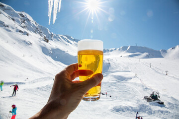 A person is holding a glass of beer with white foam in the mountains with snow on a sunny day. A...