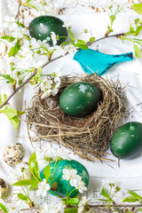 Fototapeta na wymiar Easter eggs colored with nettles in green color on table in nest and around it on white tablecloth in green wooden bird and flowering cherry branches background, Easter holiday table natural decor