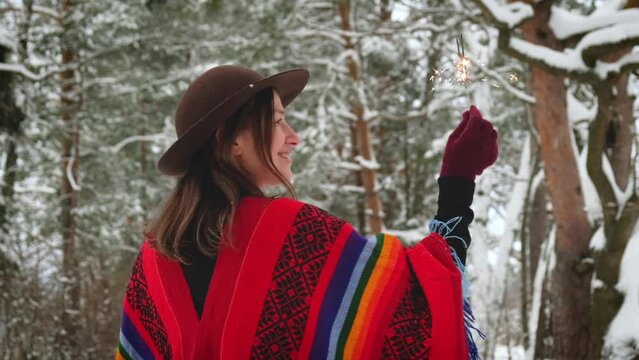 Christmas celebration outdoor, winter vacation concept. Happy positive hipster woman wearing in colorful ethnic wool poncho with sparklers in snowy forest. Person enjoy winter pleasures.