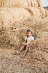 Naklejka na ściany i meble Nice young girl sitting in heap of scattered hay smiling with sly thinking eyes looking away and holding hay in hand wearing sundress. Having fun away from city on field full of golden hay.
