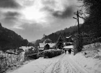 Zimowy krajobraz czarno biały, ścieżka z ludźmi oraz domy, drzewa, las, krzaki, a na niebie chmury i słońce.