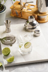 A glass with ice cubes, lime and refreshing beverage on marble board, shaker and bartender equipment
