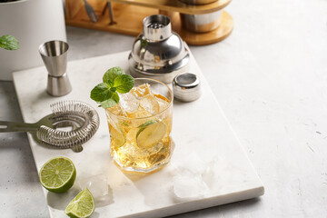 A glass with ice cubes, lime and refreshing orange beverage with mint on marble board, shaker and...