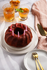 A red velvet bundt cake with birthday candles on white plate on marble surface with orange soda