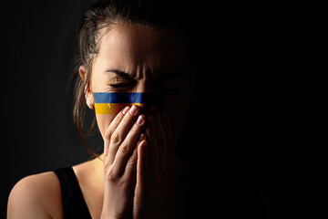 Crying hopeful sad feared frightened emotional woman with Ukraine flag on face in the dark. Stop...
