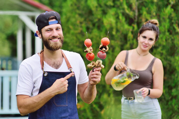 bearded handsome man smiling holding skewers with barbecue vegetables in his hands against the background of a girlfriend who pours fresh lemonade - obrazy, fototapety, plakaty