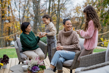 Two happy sisters with mother and grandmother sitting and drinking tea outdoors in patio in autumn.
