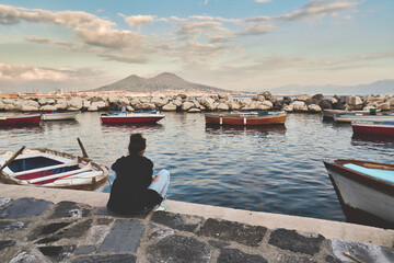 Fototapeta na wymiar Frustrated girl sitting on the Italian seashore in the middle of boats overlooking the landscape of Vesuvius
