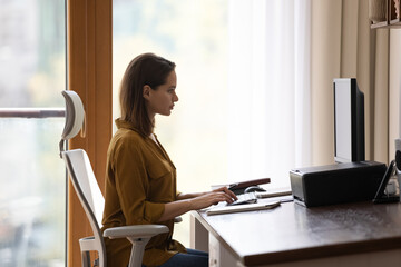Side profile view attractive young 30s serious woman sit at desk working on computer, looking at pc...