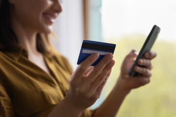 Close up young woman shopper purchasing on-line, holds debit card and cell phone makes order, instant payment, send funds using virtual secure e-services, spend money on internet. E-commerce concept