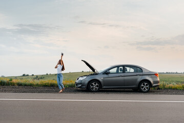 Fototapeta na wymiar A young girl stands near a broken car in the middle of the highway during sunset and tries to call for help on the phone and start the car. Waiting for help. Car service. Car breakdown on the road.