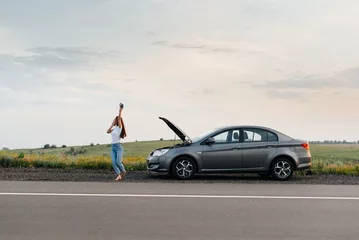 Fotobehang A young girl stands near a broken car in the middle of the highway during sunset and tries to call for help on the phone and start the car. Waiting for help. Car service. Car breakdown on the road. © Andrii