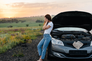 A frustrated young girl stands near a broken-down car in the middle of the highway during sunset. Breakdown and repair of the car. Waiting for help. Car service. Car breakdown on road.