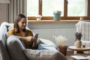 Beautiful young 30s woman holds smartphone smile read pleasant message, enjoy new mobile application relaxing alone on sofa. Buying goods e-services on internet, making order, do e-shopping at home