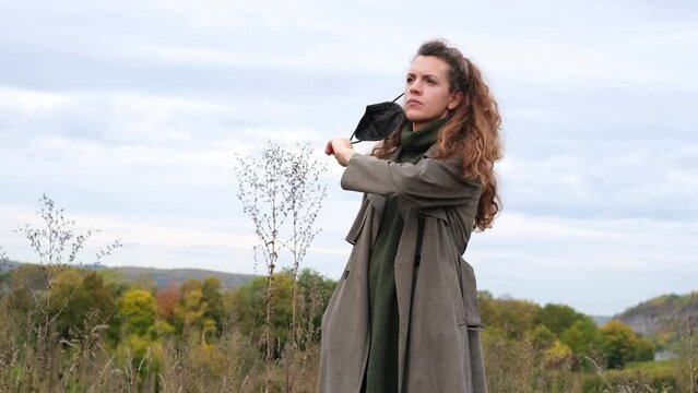 Portrait of a beautiful, stylish hipster woman standing in nature, taking off a black ffp2 respirator mask and throwing it away. Autumn landscape, fresh air, freedom and recreation of modern people.