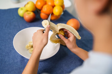 Close up on hands of unknown caucasian woman cutting banana by the table at home - back view top...