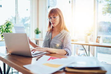 Fototapeta premium Beautiful skilled female freelancer analyzing data from online reports on laptop computer, prosperous business woman use eyewear for vision protection check exchange news work remotely in cafeteria .