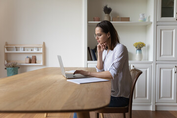 Young woman in glasses sit at table looks at laptop screen makes research use internet, web surfing...