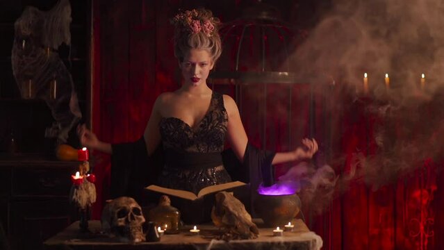 Halloween witch with cauldron and magic book. Beautiful woman flowers in hair conjuring making witchcraft. Take off black cloak Enchantress prepare love potion use magic spell Slow motion. Handheld