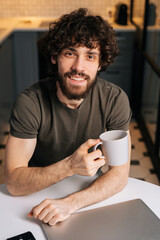 High-angle view of smiling bearded young business man holding in hand cup with morning coffee sitting at table with closed laptop computer, looking at camera, in kitchen with modern interior.