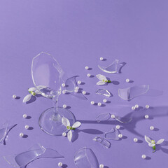 Party composition made with pieces of broken wine glass,  white pearls and snowdrop flowers with sunshine shadows on purple background. Creative spring holiday concept.
