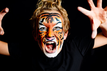 A growling boy with his face painted in the striking colors of a tiger, isolated on a black studio...
