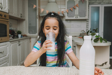 Cute little girl drinks milk at the table in the kitchen. the child is holding a bottle of milk....