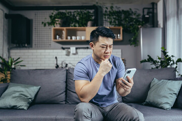 Obraz premium Concerned man reading news online looking at phone screen, Asian sitting on sofa at home, serious and sad