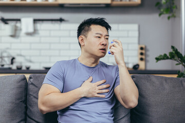 A man with asthma, an Asian man at home sitting on the couch breathing in an inhaler - obrazy, fototapety, plakaty