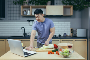 male chef cooks at home and teaches online, Asian cuts vegetables in the kitchen, uses laptop for online cooking classes