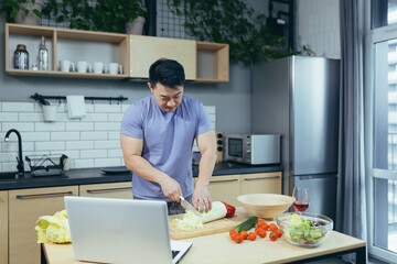 male chef cooks at home and teaches online, Asian cuts vegetables in the kitchen, uses laptop for online cooking classes