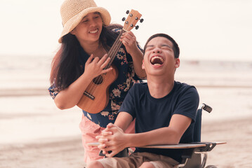 Young man with disability and parent or volunteer or caregiver smiling and singing, playing ukulele on the beach,Vacation on holiday with family activity and natural therapy and mental health concept.