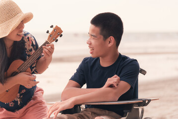 Young man with disability and parent or volunteer or caregiver smiling and singing, playing ukulele on the beach,Vacation on holiday with family activity and natural therapy and mental health concept.