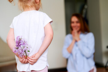 Mothers Day. A little preschool girl holds a bouquet of lilac flowers behind her back and wants to give it to her mom