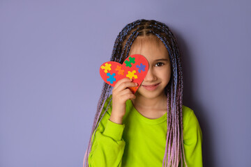 A girl with blue-purple Afro pigtails in a light green top holds a heart with puzzle pieces in her hands as a sign of solidarity with people suffering from autism spectrum disorder. World Autism day - obrazy, fototapety, plakaty
