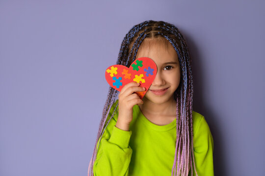 A girl with blue-purple Afro pigtails in a light green top holds a heart with puzzle pieces in her hands as a sign of solidarity with people suffering from autism spectrum disorder. World Autism day