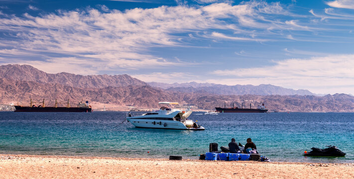 Sunny day on sandy beach of the Red Sea with pleasure yachts, motorboats and cargo ships near Eilat - famous tourist resort and recreational Israeli city 