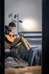 A young man plays the acoustic guitar in his room at home.
